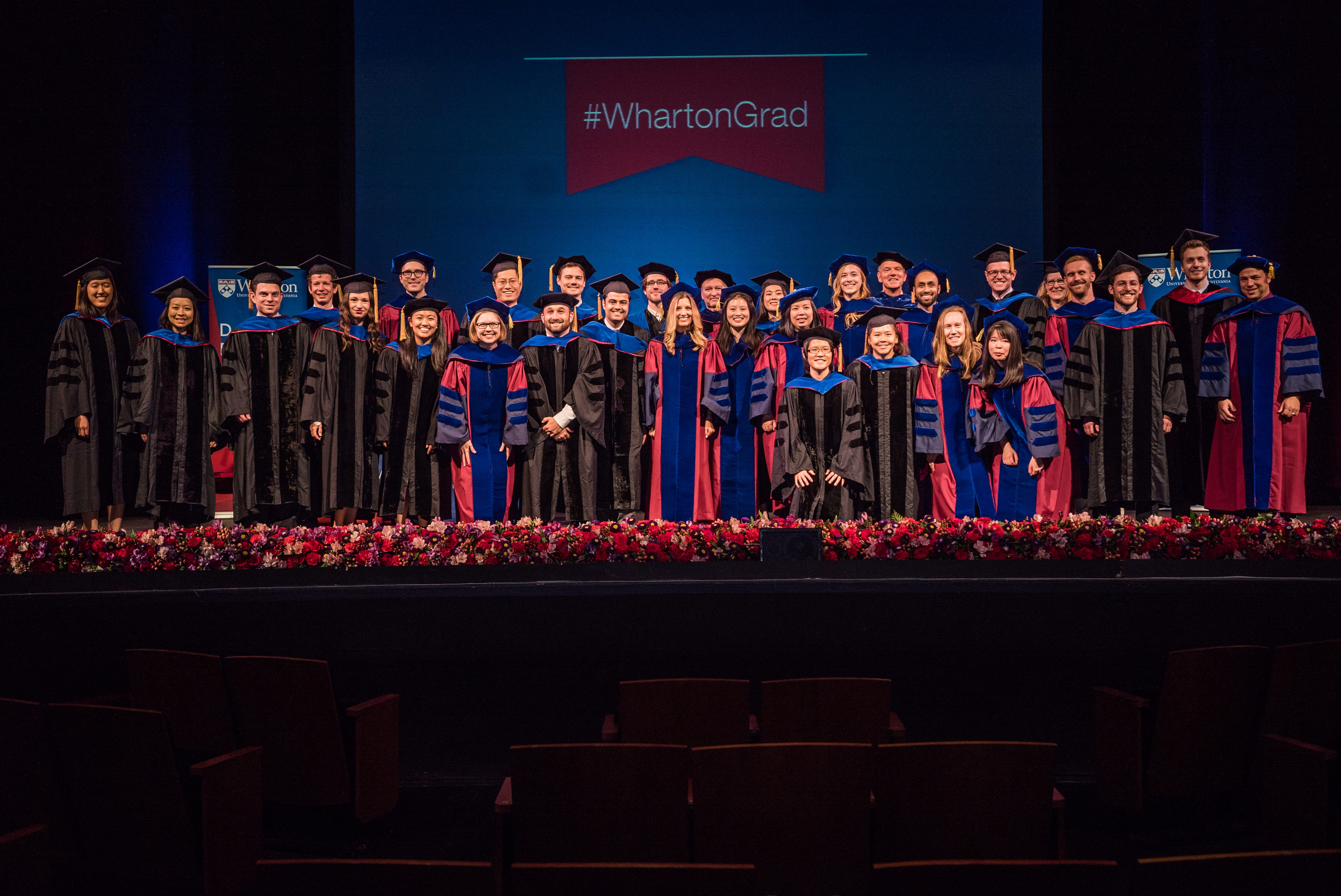 A group of people in graduation robes and caps standing on a stage with a banner reading "#WhartonGrad."